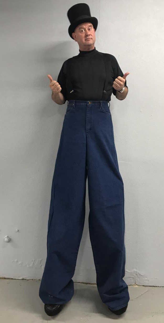 A man is standing on stilts wearing a top hat and jeans.