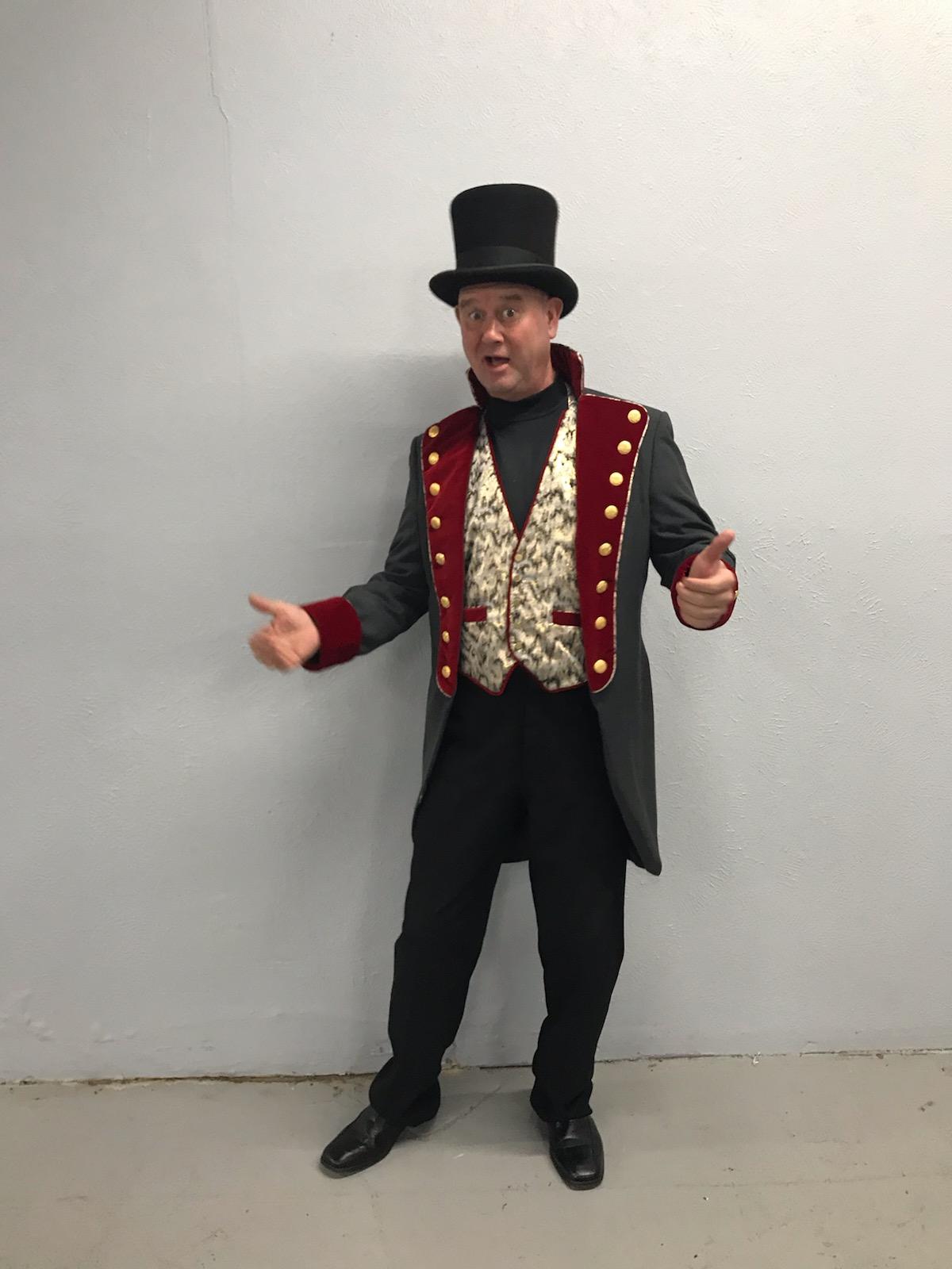 A man in a top hat and coat is standing in front of a white wall.