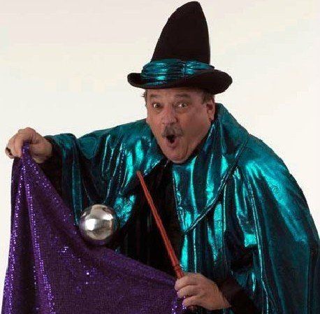 A man in a witch costume is holding a purple cloth and a wand