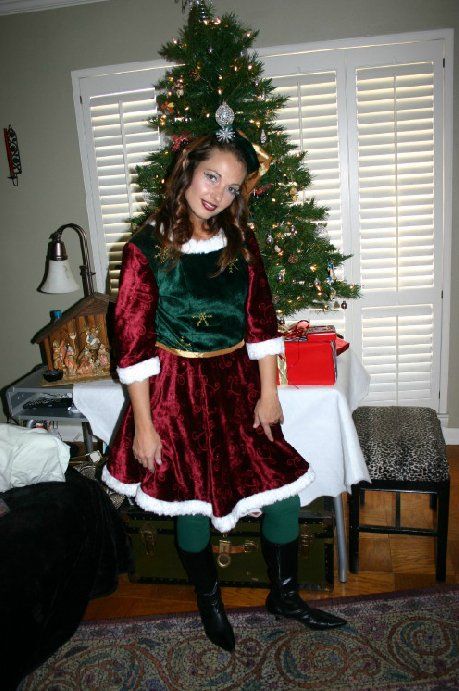 A woman dressed as an elf is standing in front of a christmas tree.