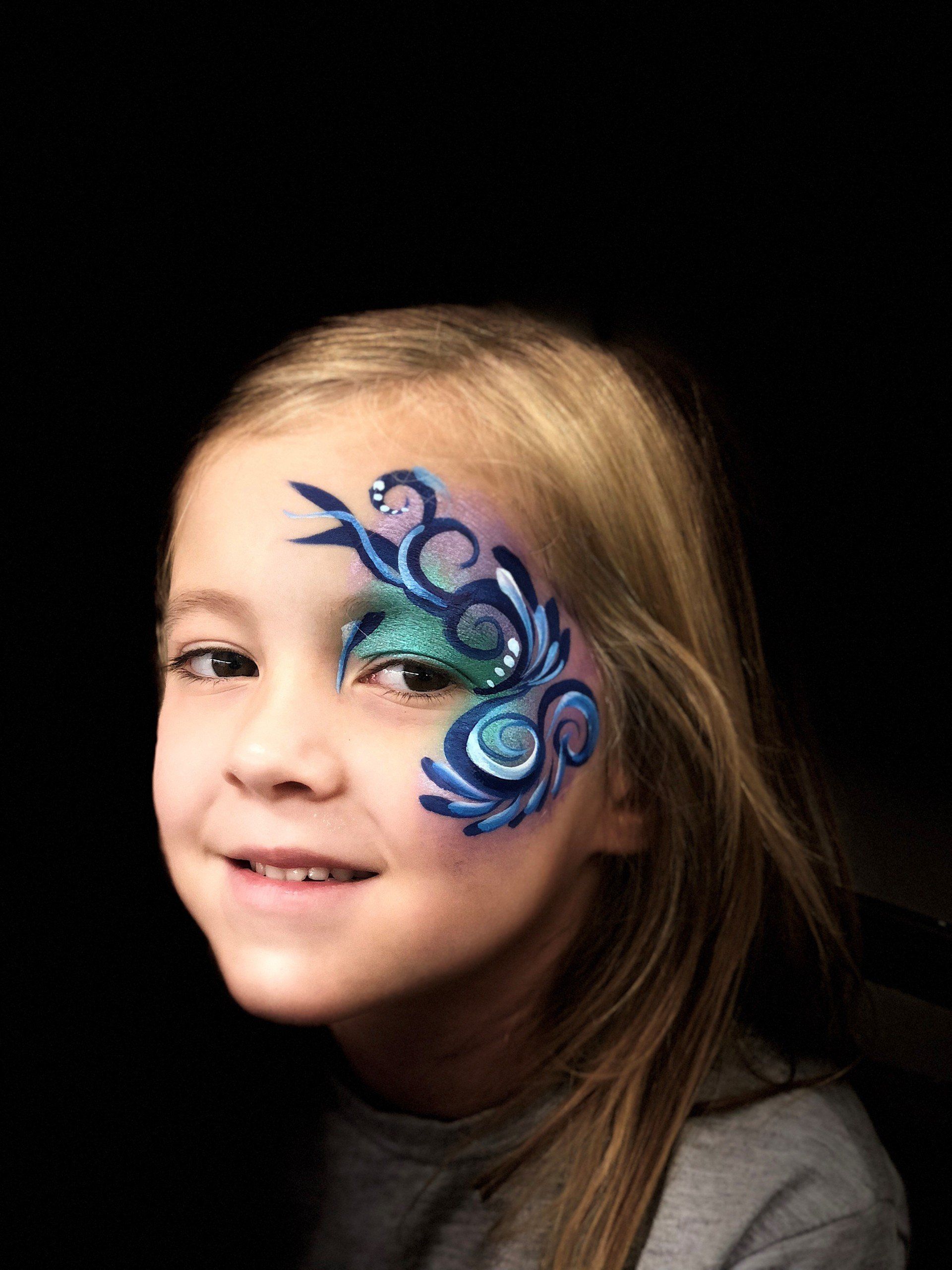 A little girl with a peacock feather painted on her face.