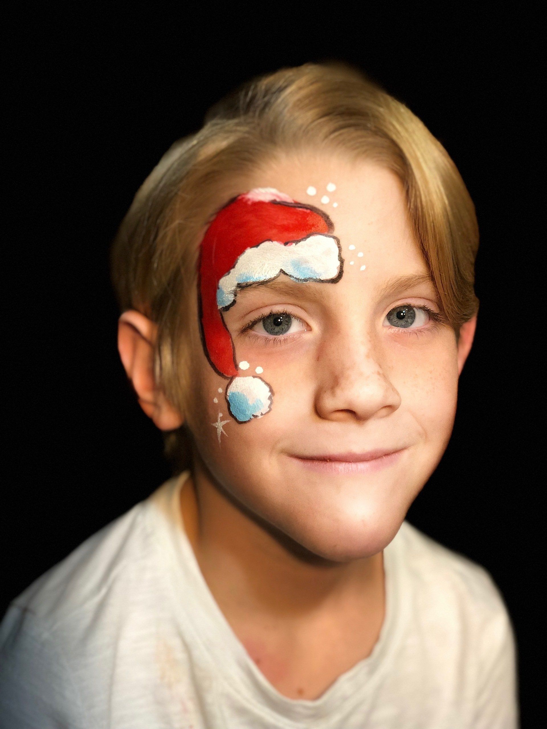 A young boy has a santa hat painted on his face.