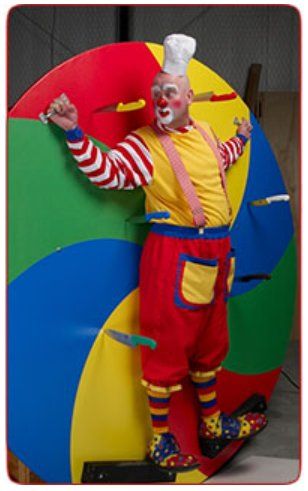 A clown is standing in front of a spinning wheel with knives sticking out of it