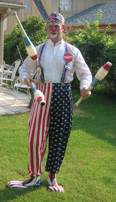 A clown is standing in the grass holding a juggling stick.