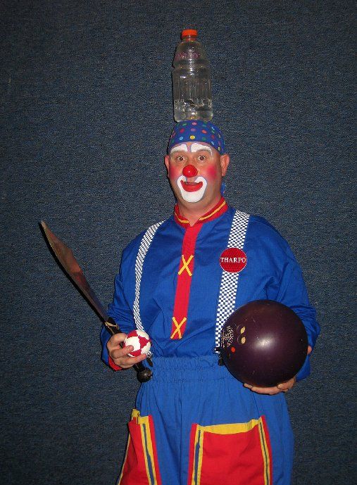A clown is holding a bowling ball and a bottle on his head