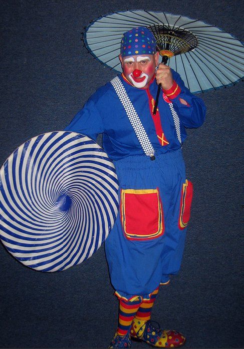 A clown in a blue outfit is holding an umbrella and an optical illusion