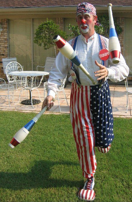 A man dressed as a clown is juggling clubs in a yard.