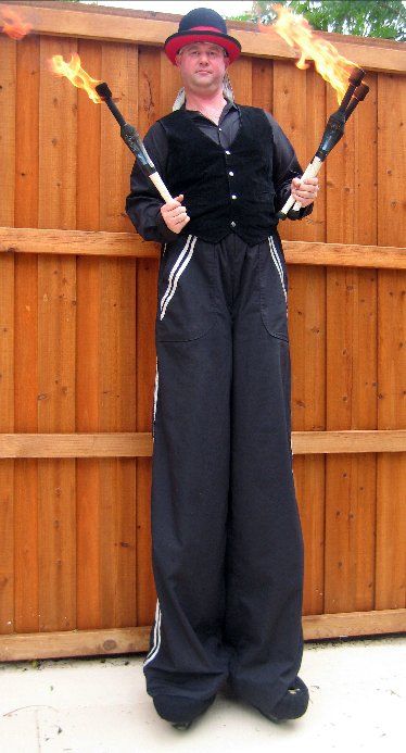 A man on stilts is holding two fire torches in front of a wooden fence.