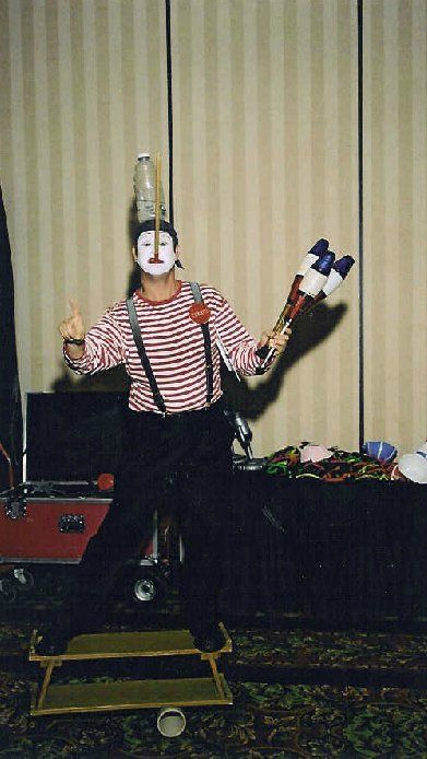 A man in a clown costume is juggling a bottle on his head.