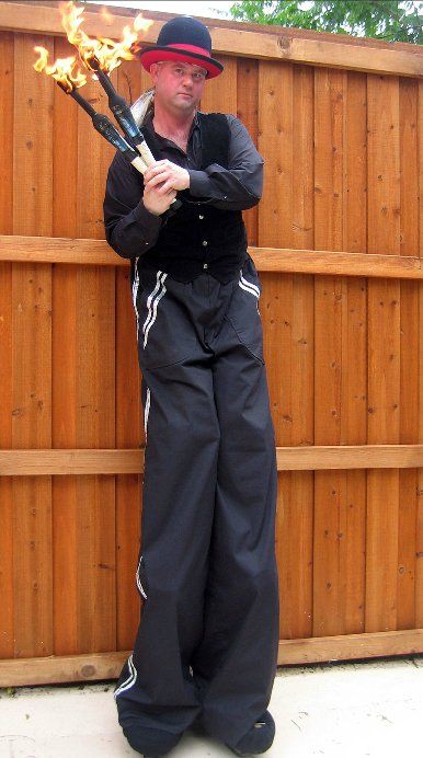 A man on stilts is holding a torch in front of a wooden fence.