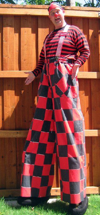A man in a clown costume is standing on stilts in front of a wooden fence.