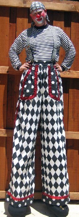 A man in a clown costume is standing on stilts in front of a wooden fence.