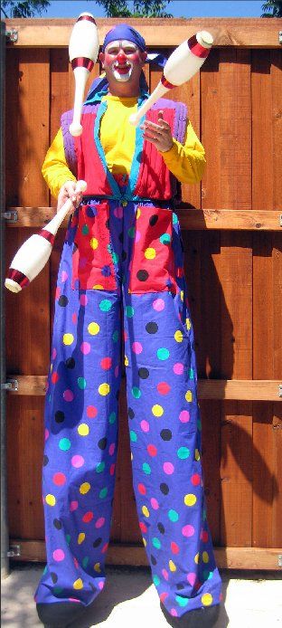 A clown on stilts holding juggling pins in front of a wooden fence