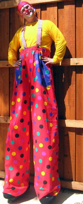 A clown on stilts is standing in front of a wooden fence.