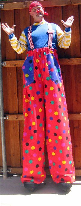 A clown on stilts is standing in front of a wooden fence