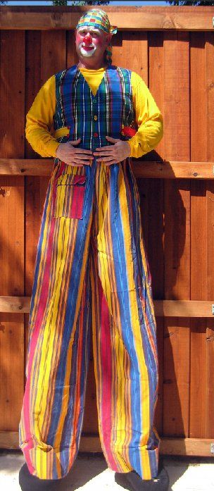 A clown on stilts is standing in front of a wooden fence.