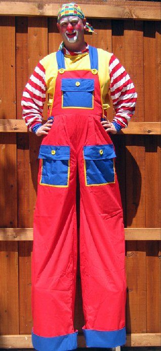 A clown on stilts is standing in front of a wooden fence.