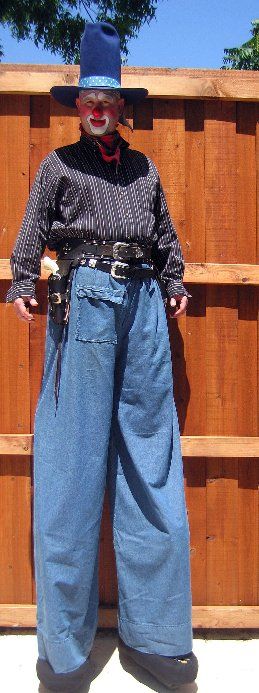 A clown is standing on stilts in front of a wooden fence.