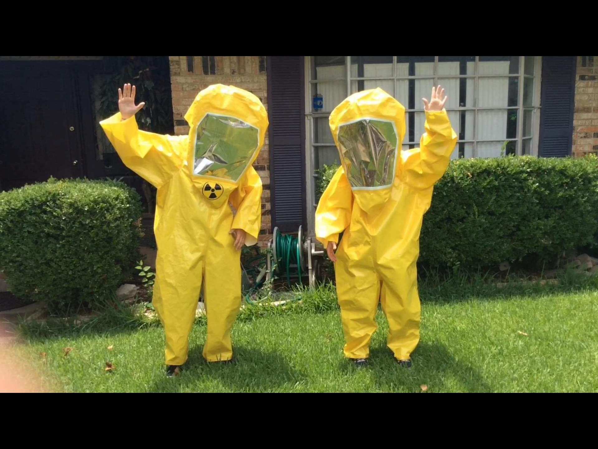 Two children wearing yellow protective suits are standing in the grass.