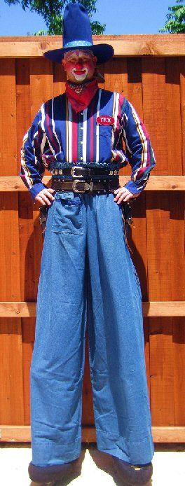 A clown on stilts is standing in front of a wooden fence