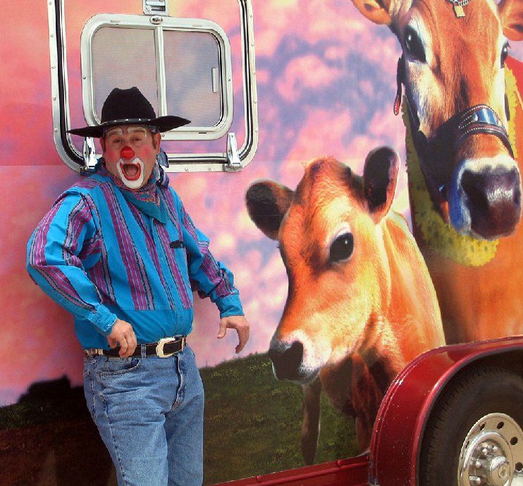 A man dressed as a clown stands in front of a cow mural