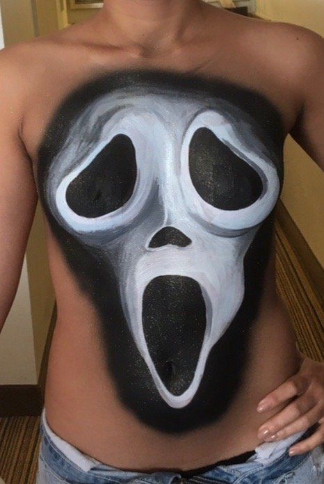 A woman has a scream mask painted on her chest.