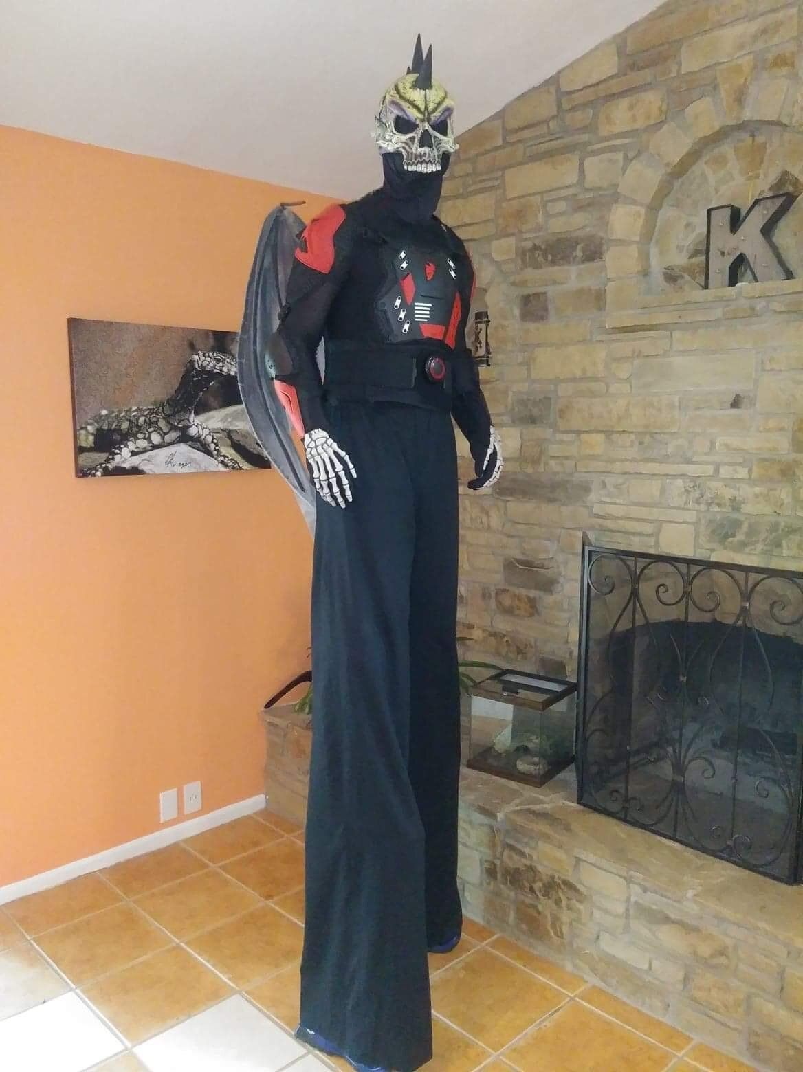A man in a skeleton costume is standing on stilts in front of a fireplace.