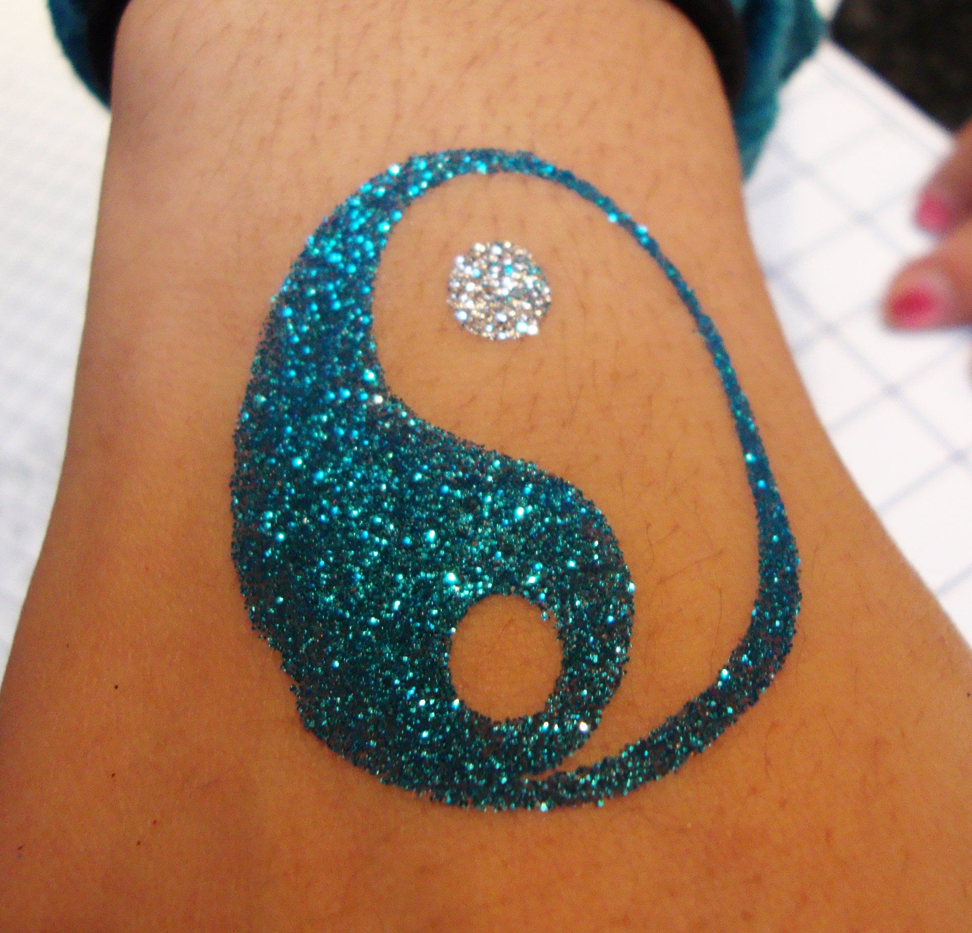 A person has a blue glitter tattoo of a yin yang symbol on their wrist.