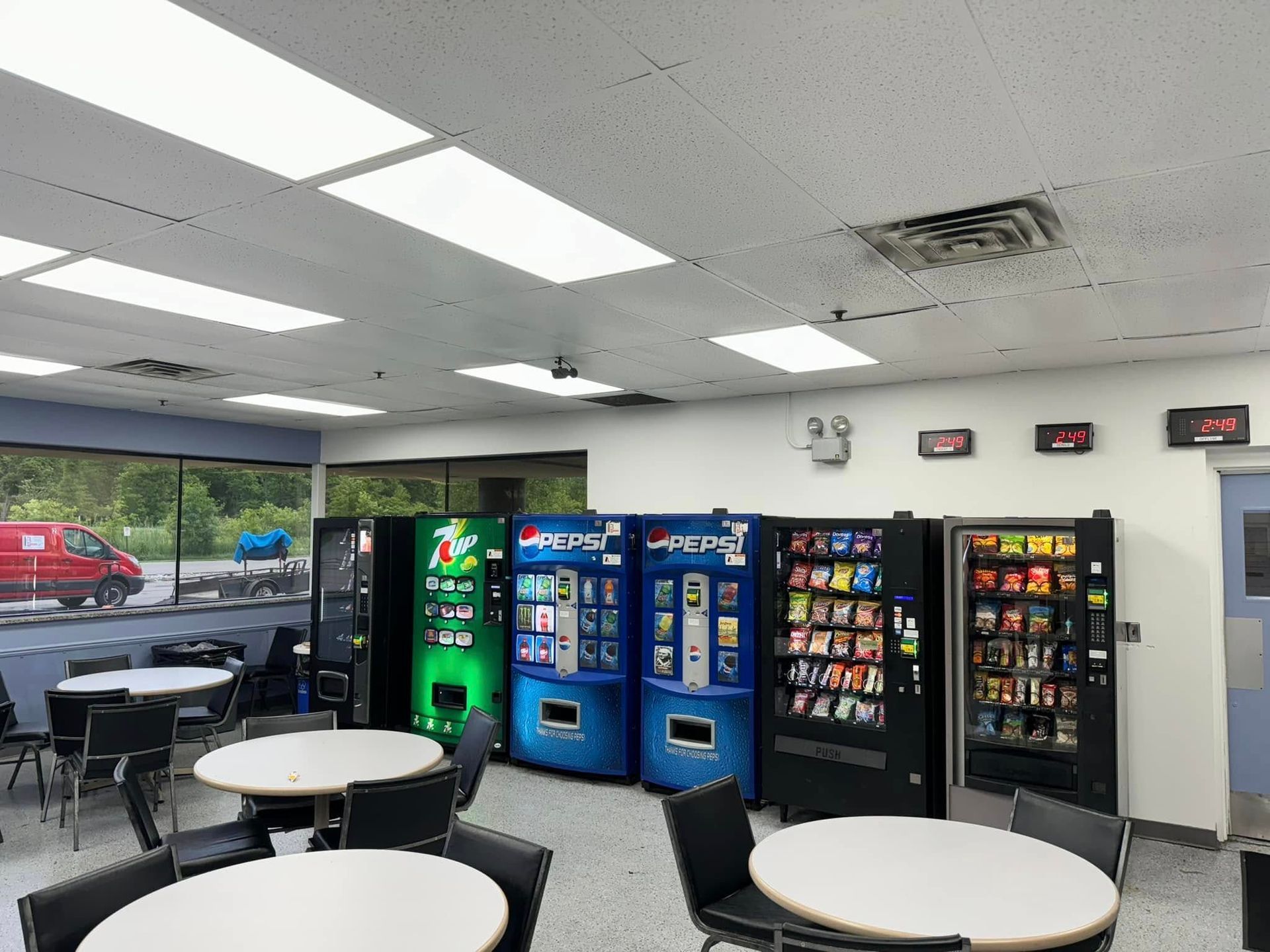 Interior of a break room with vending machines, tables, and chairs. Fluorescent lighting illuminates the space.