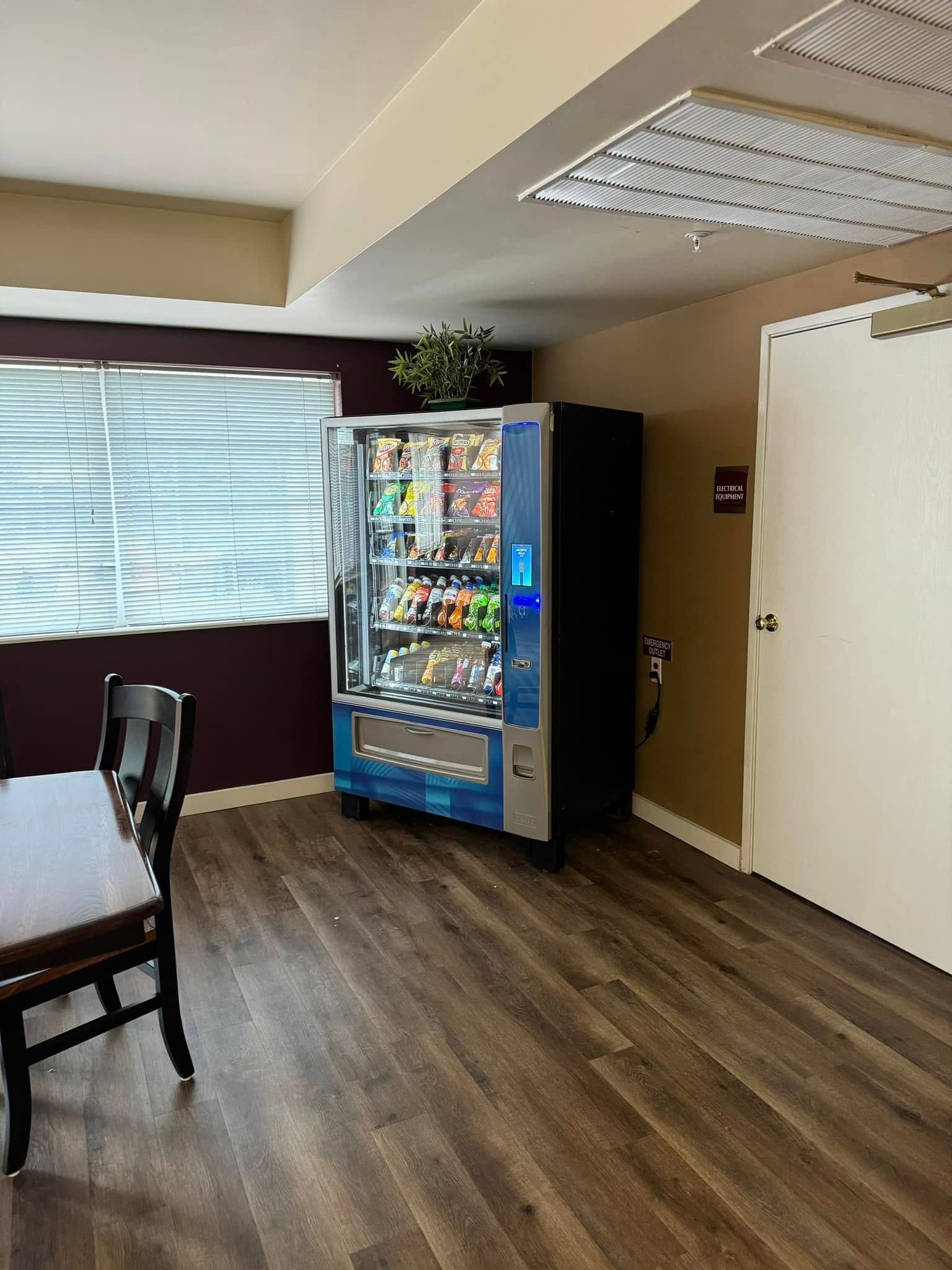 Vending machine in a room with a window, chair, and door, featuring various snacks and drinks.