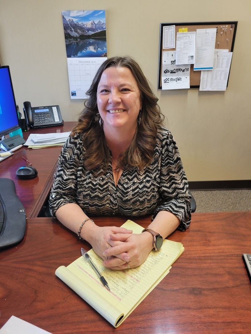 A woman is sitting at a desk with her hands folded and smiling.