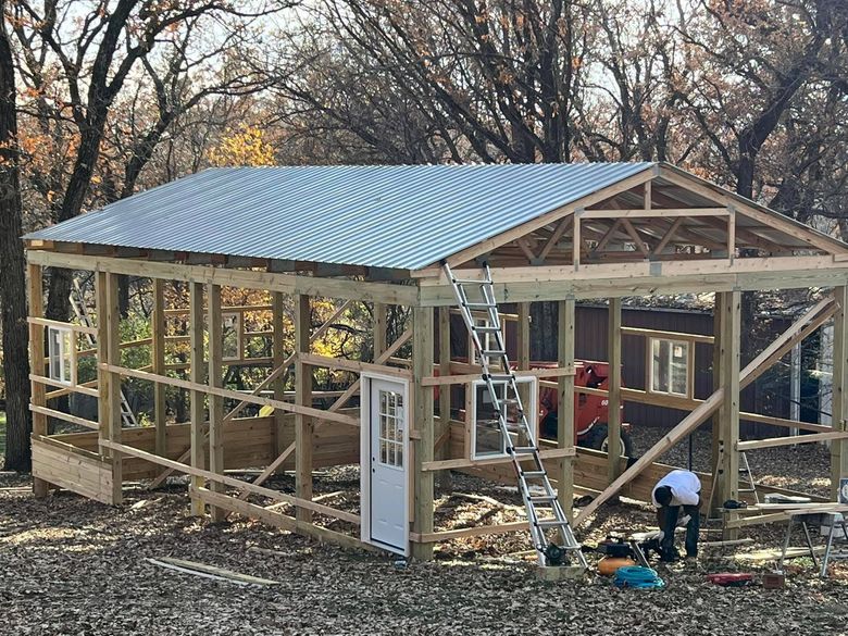 A partially constructed wooden frame building with a metal roof, located in a wooded area with a person working nearby.