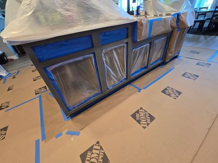 A kitchen island and cabinets partially covered in blue painter's tape and plastic sheets for a painting project.