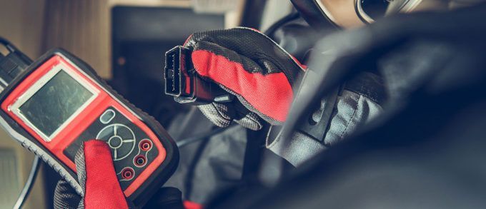 Gloved hands holding a red handheld device with a small screen and buttons in a blurred indoor setting