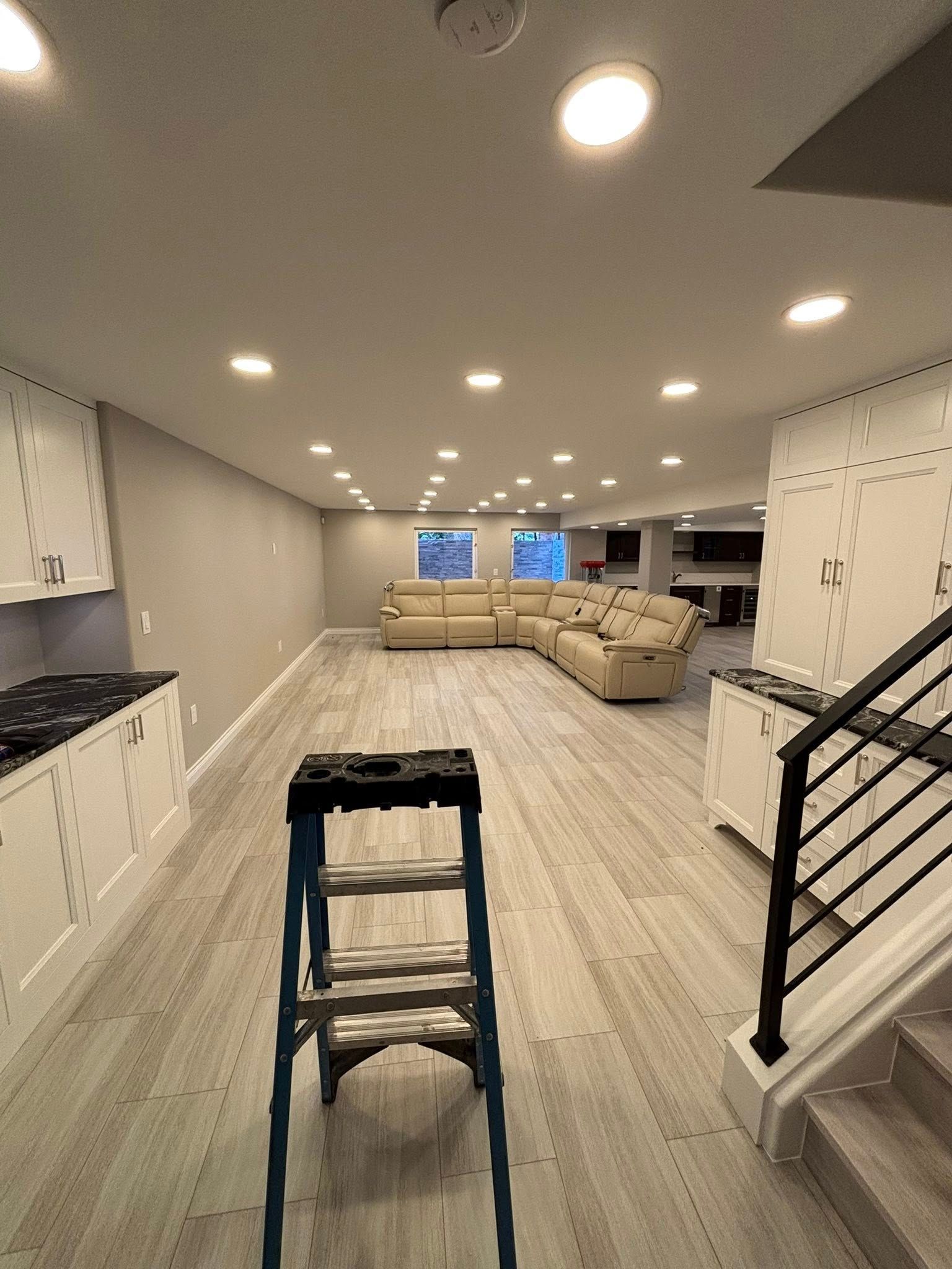 A ladder stands in a renovated room with light wood floors, recessed lights, and a sectional sofa.