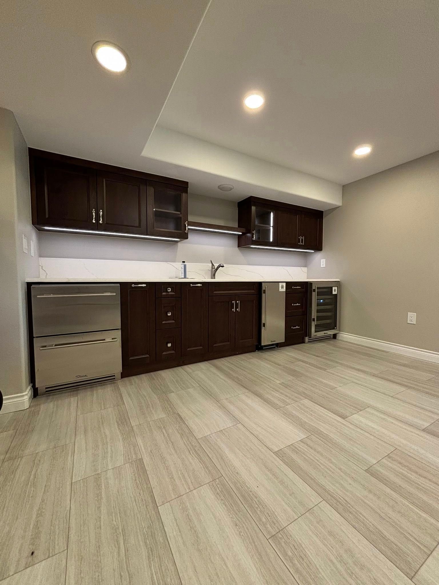 Dark brown basement bar with stainless steel appliances and light wood-look flooring.