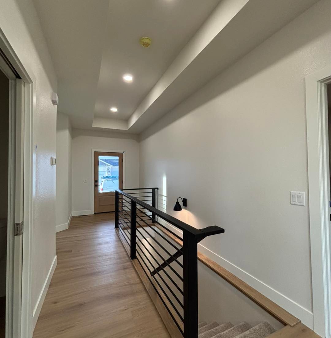 Hallway with light wood floors, white walls, and black railing. A door is at the end.