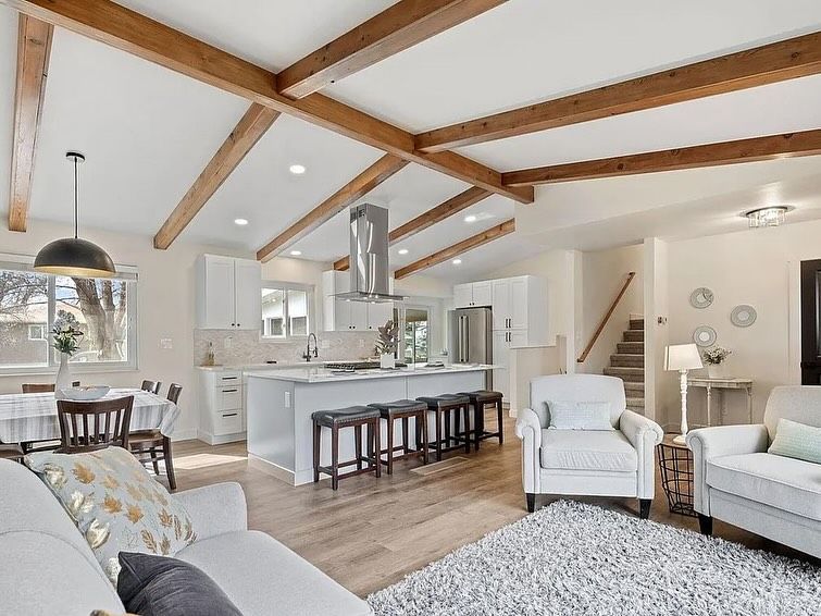 Open-plan living area with white cabinets, island, wood beams, seating, dining table, stairs, and a light gray rug.
