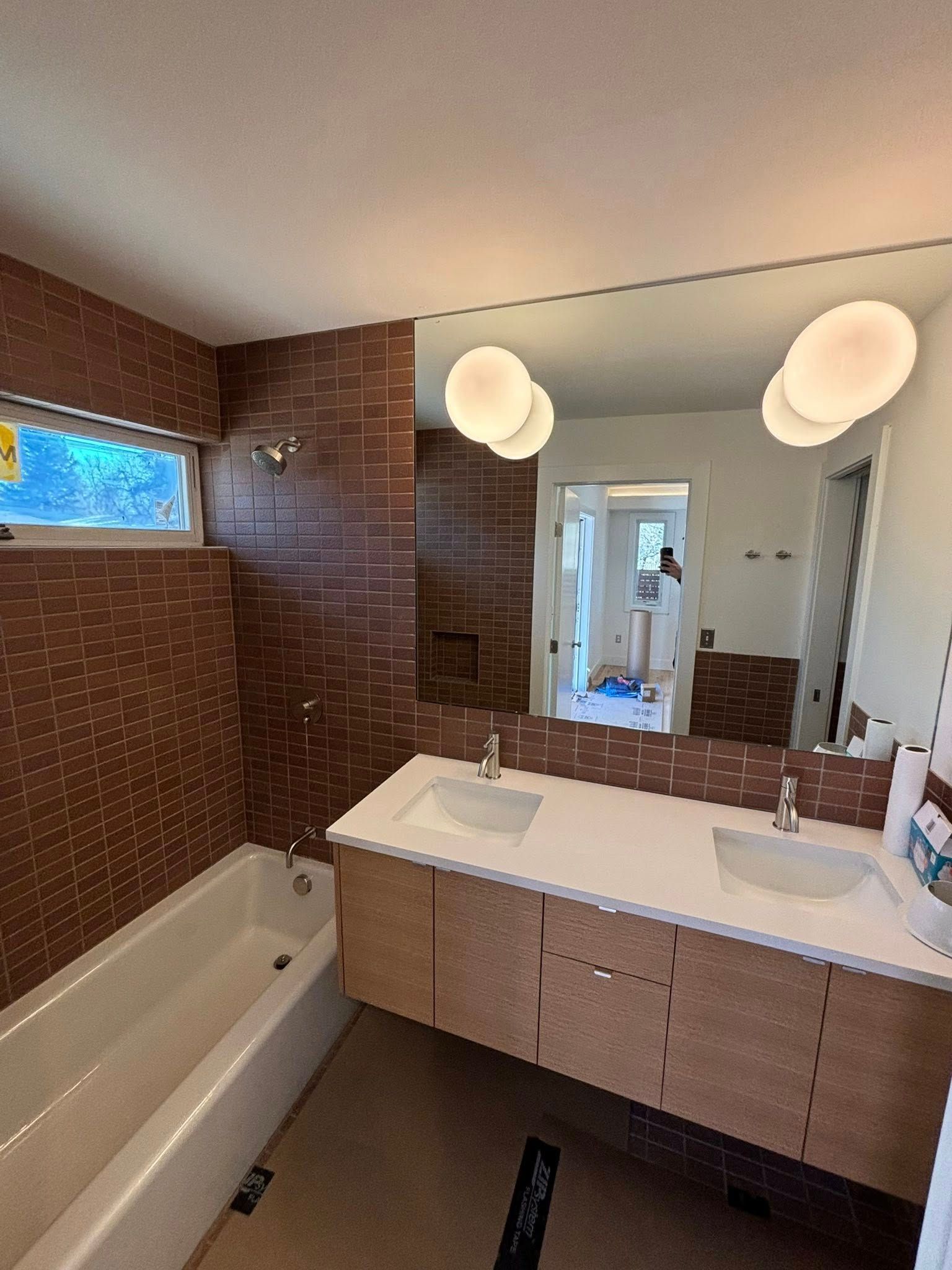 Bathroom with a rectangular tub, double vanity, and a large mirror. Brown tiled walls, two pendant lights.