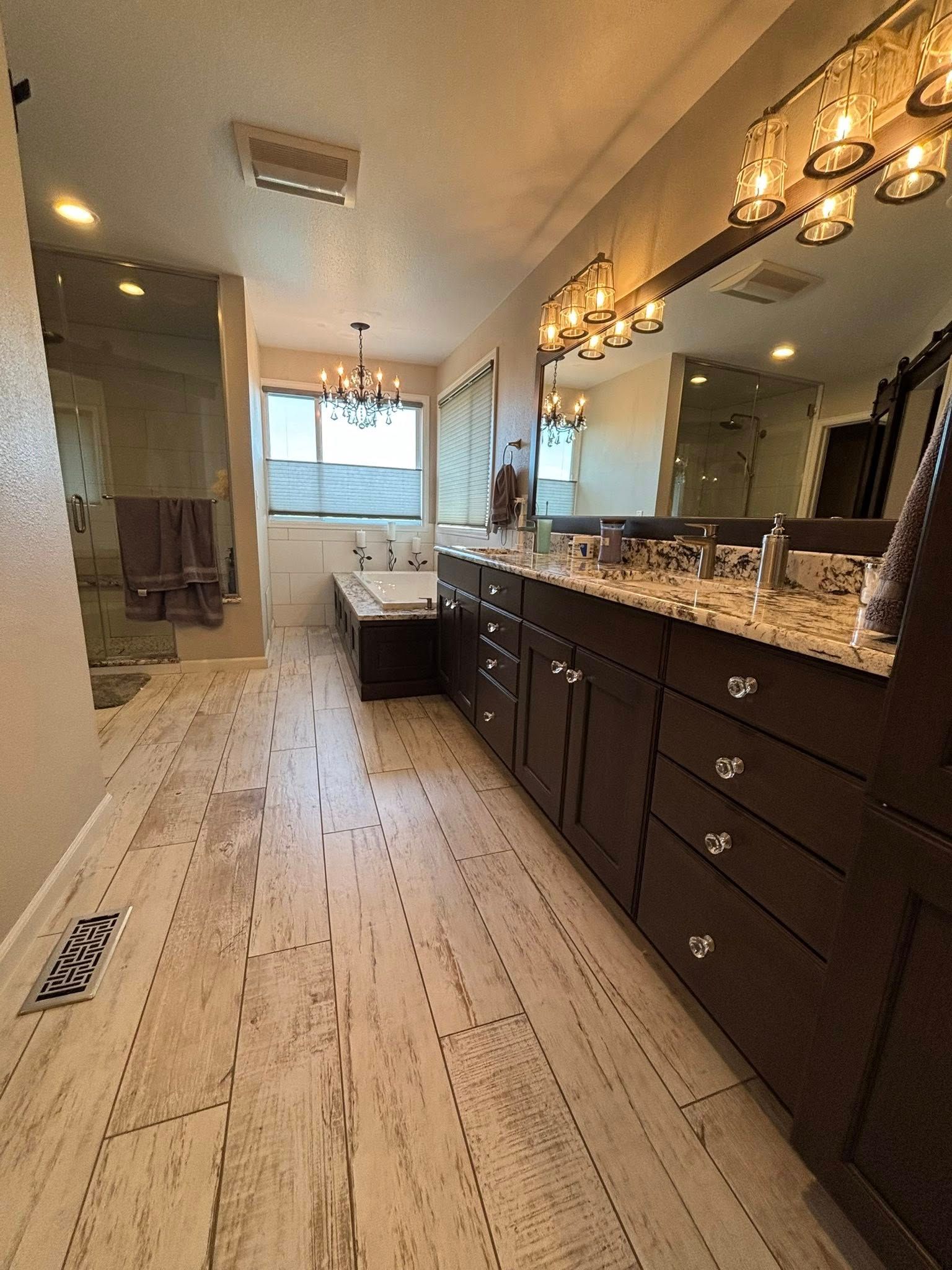 Spacious bathroom with wood-look tile flooring, dark wood vanity, glass-enclosed shower, and a soaking tub.