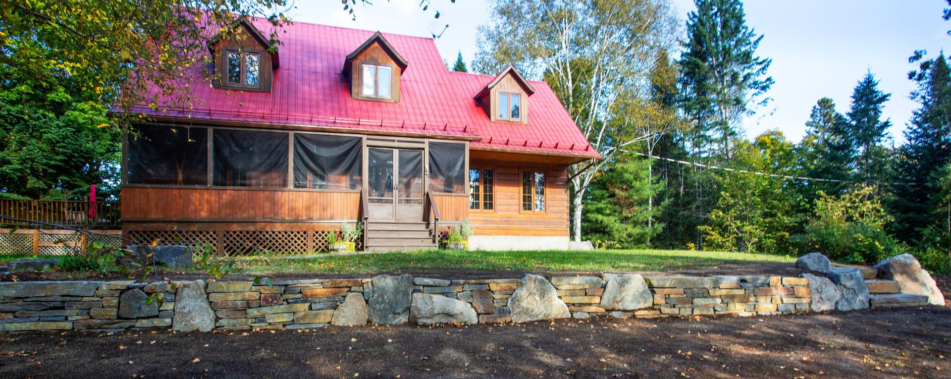 Une grande maison en bois avec un toit rouge est entourée d'arbres et d'un mur de pierre.
