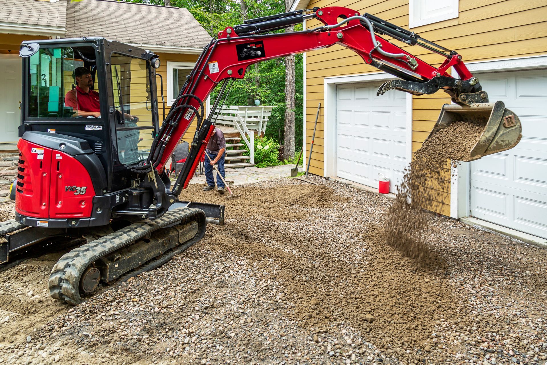 Une excavatrice rouge creuse une allée de gravier devant une maison.