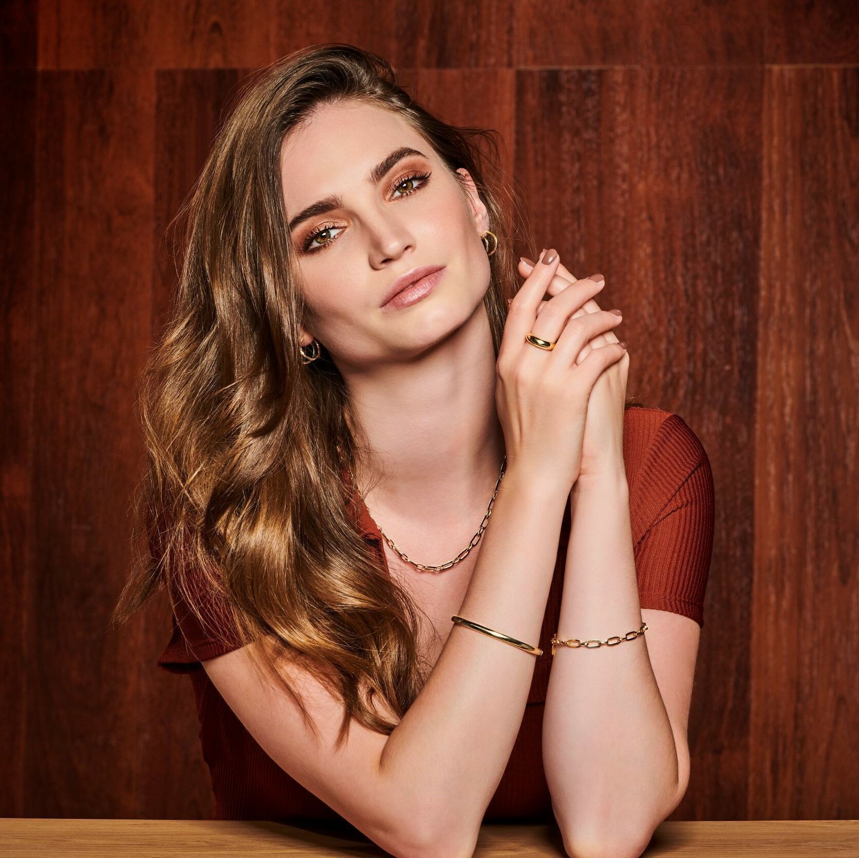 Een vrouw met een ketting en een armband zit aan een tafel met haar handen gevouwen.