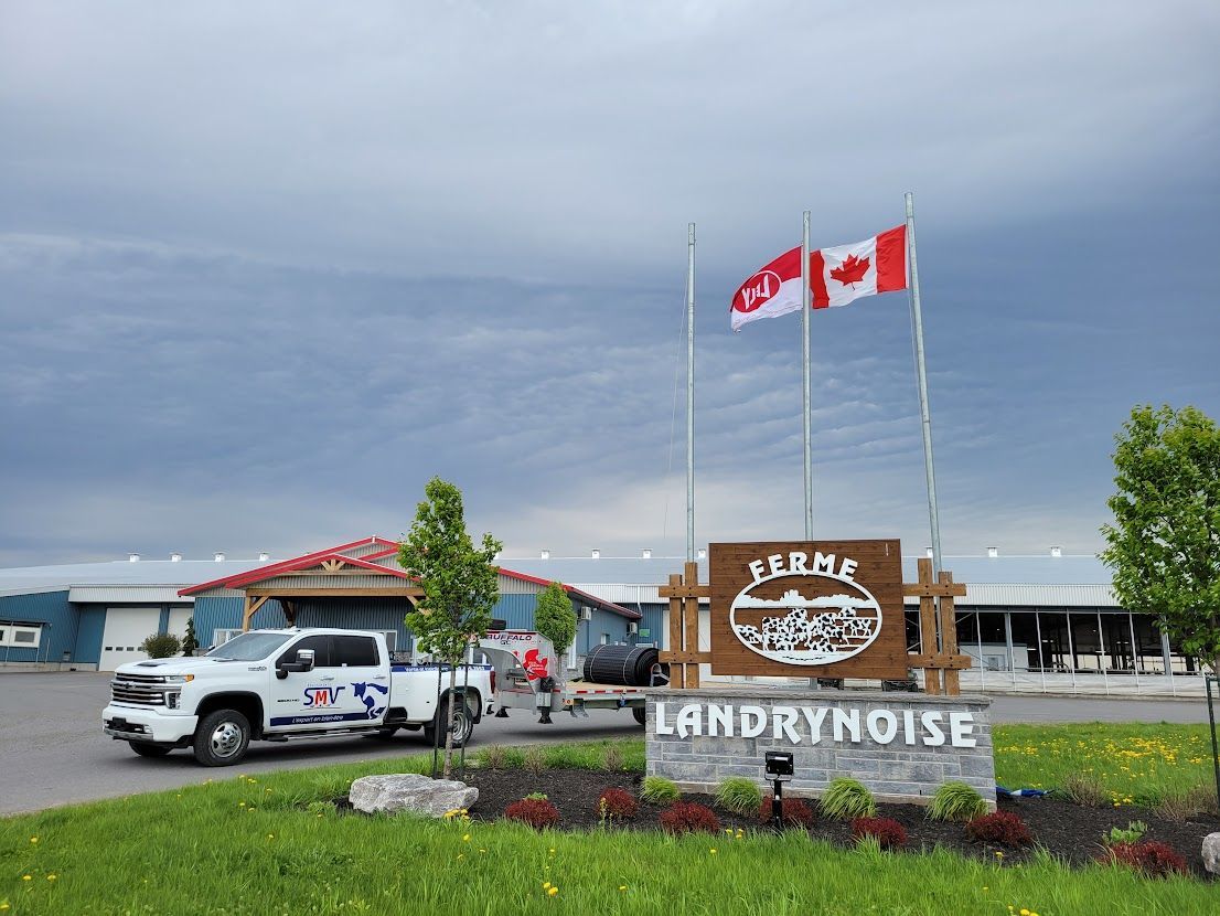 Panneau pour la Ferme Landry Ferme bruyante devant des bâtiments avec deux drapeaux canadiens et une camionnette blanche avec une remorque.