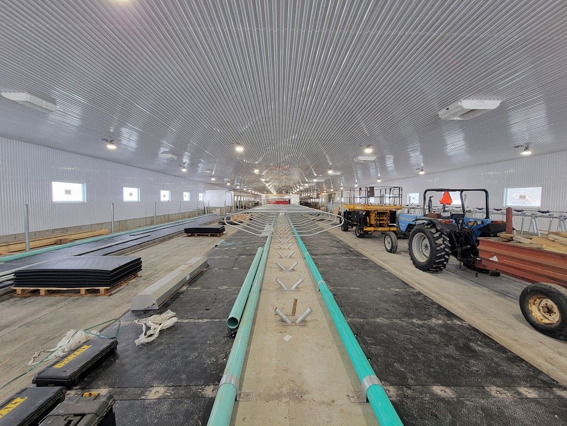 Vue intérieure d'une grande grange en construction avec sol en béton, tracteur bleu et matériaux de construction. Le plafond est à motifs.