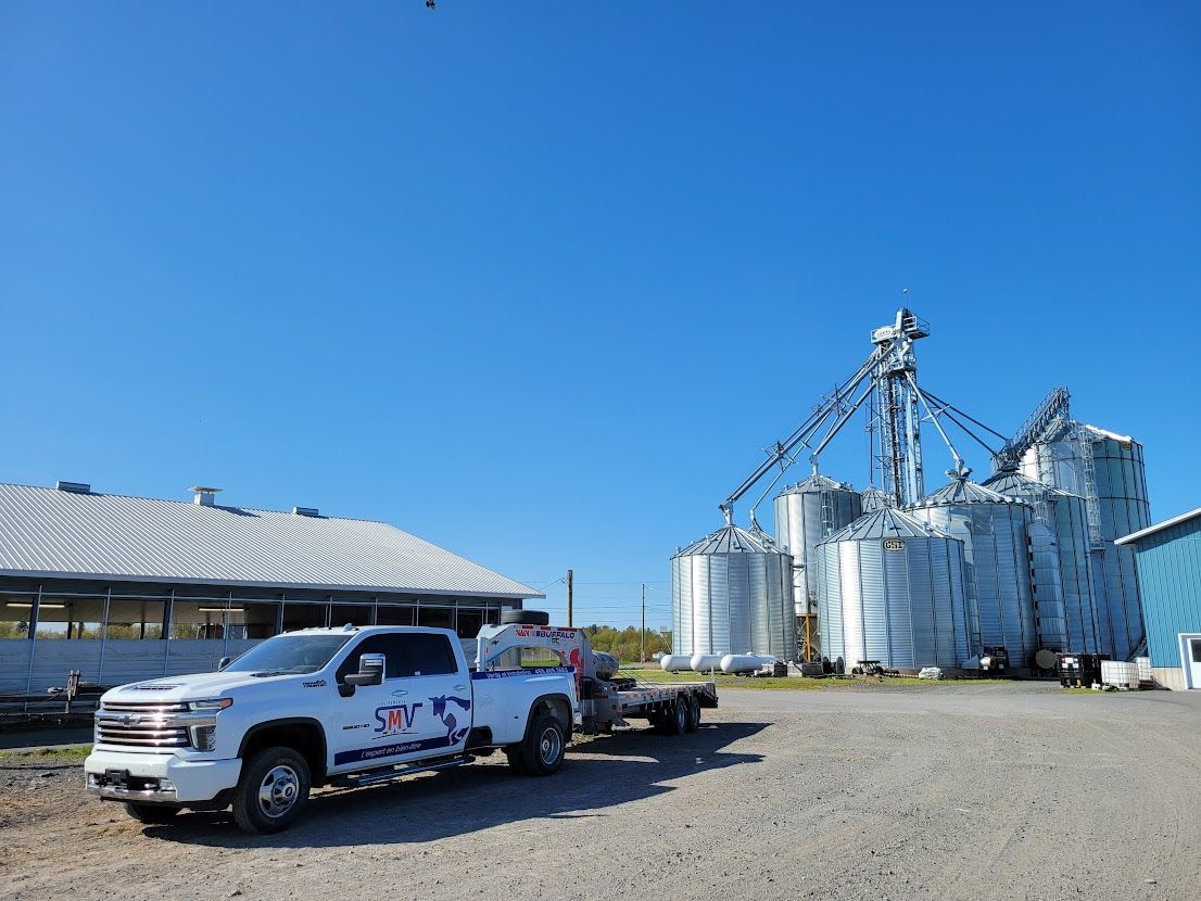 Camion blanc avec une remorque garée devant des silos à grains argentés et un ciel bleu.