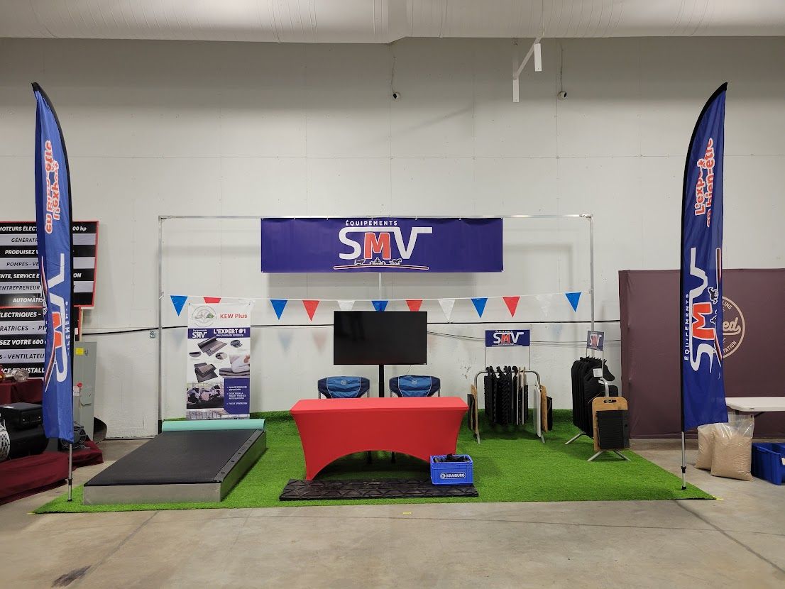 Un stand événementiel avec une table rouge, un téléviseur et un sol en gazon synthétique. Des drapeaux avec logos encadrent l'affichage, surmonté d'une bannière.