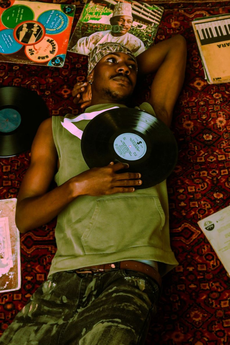 Person lying on a patterned rug, holding a vinyl record, surrounded by albums and a keyboard.