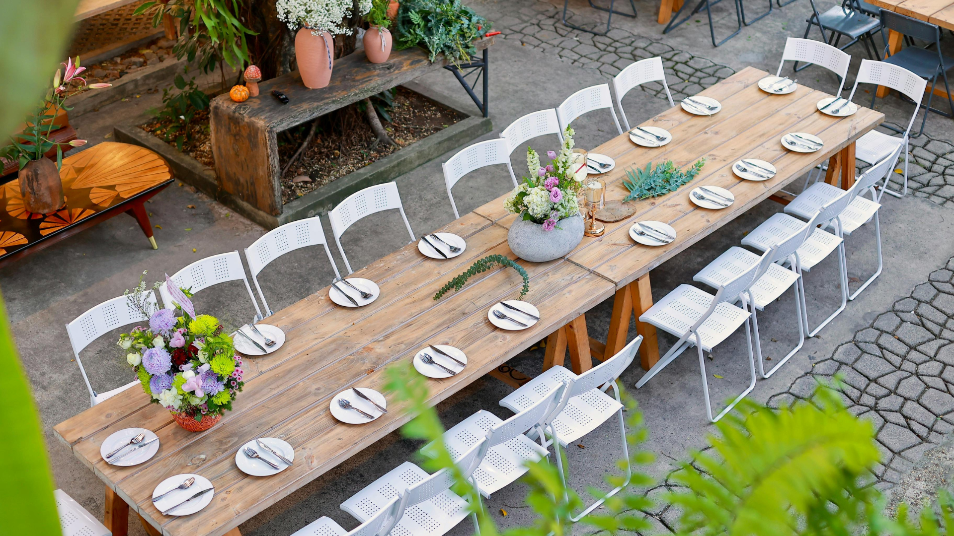 Outdoor long wooden table set for a meal with white chairs, plates, and floral centerpieces