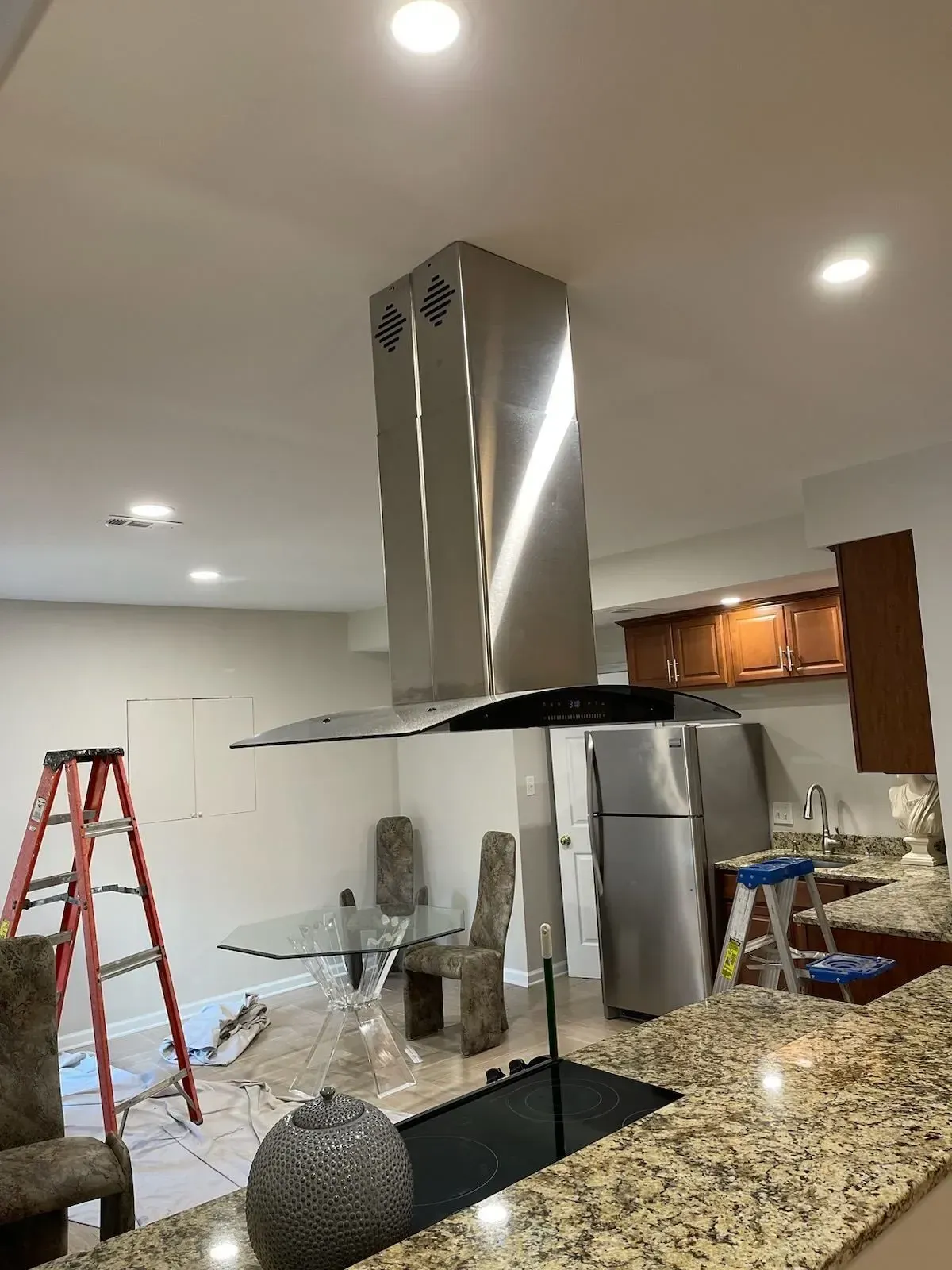 Stainless steel range hood over a kitchen island with a granite countertop, in a room with a table, chairs, and refrigerator.